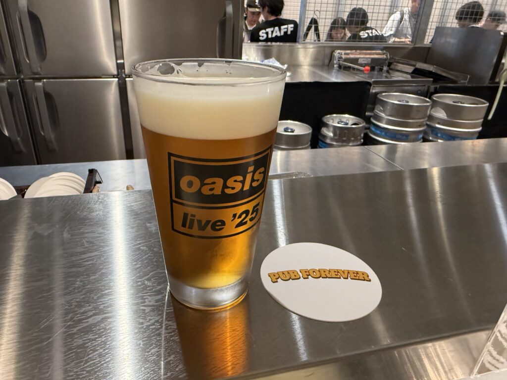 ステンレス製のバーカウンターに置かれた、Oasis live '25のロゴ入りグラスに入ったビールと、「PUB FOREVER」と書かれた白いコースター。