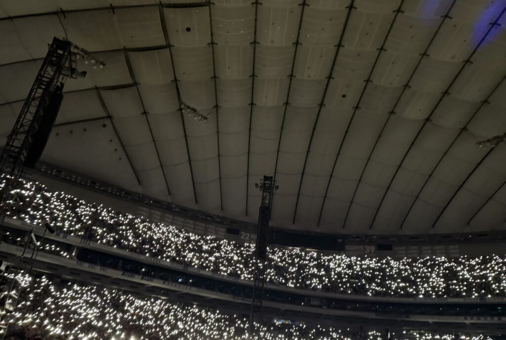 The ceiling of a large domed stadium, with the stands filled with a crowd holding up their smartphone flashlights, creating a sea of white lights.