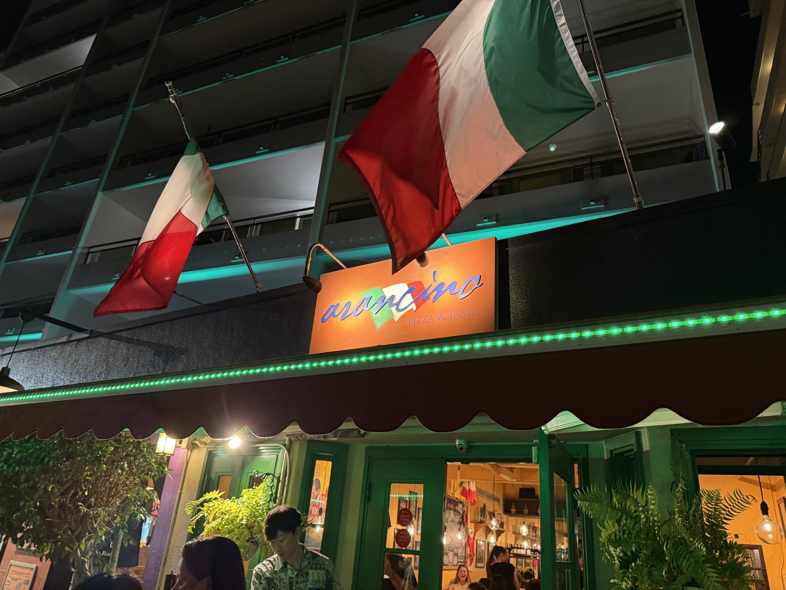 The exterior of Arancino, an Italian restaurant, at night. The entrance is decorated with a glowing orange sign, green lights, and two Italian flags.