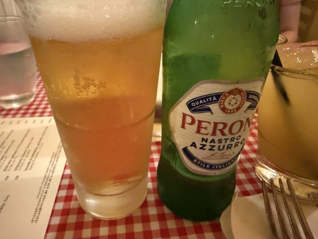 A bottle of Peroni beer and a tall, foamy glass of beer on a red and white checkered tablecloth at an Italian restaurant.