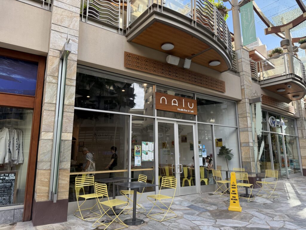 The exterior of NALU Health Bar & Café at Waikiki Beach Walk, featuring its bright yellow chairs on the stone-paved patio.