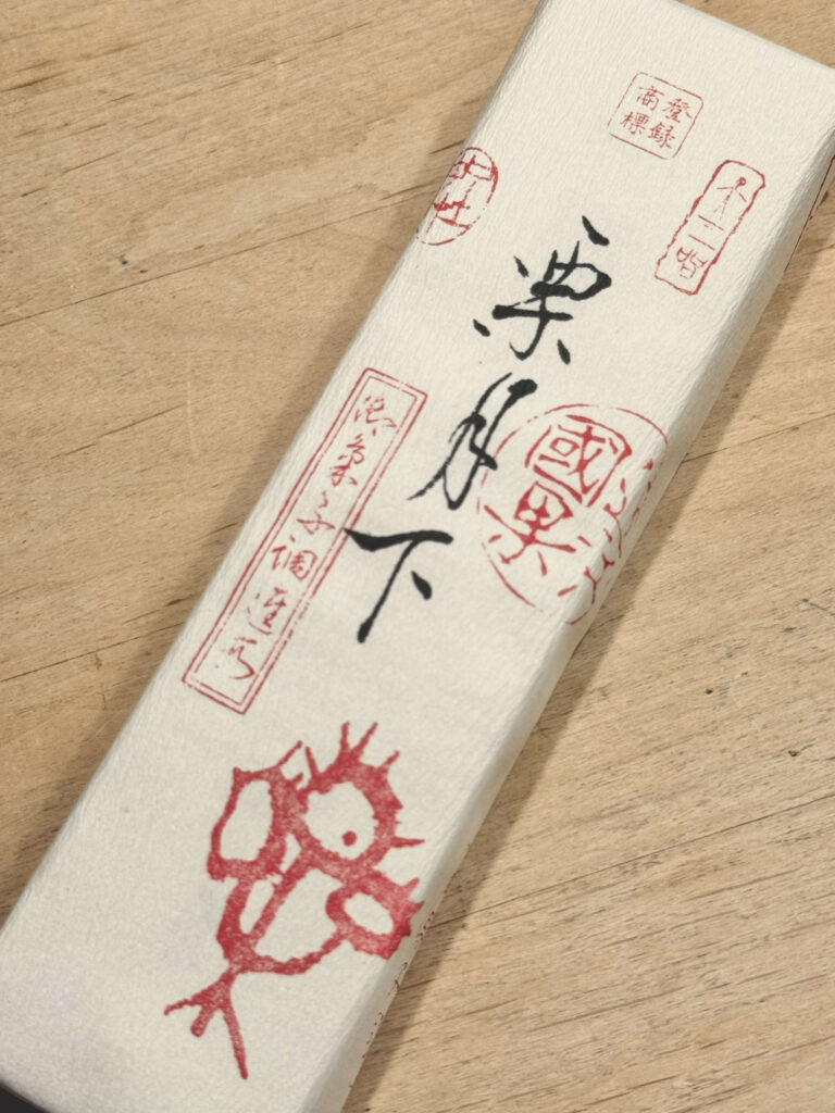 木製のテーブルの上に置かれた和菓子「栗月下」の白い箱。箱には黒い毛筆の文字と、龍の絵柄を含む複数の赤い印が押されている。