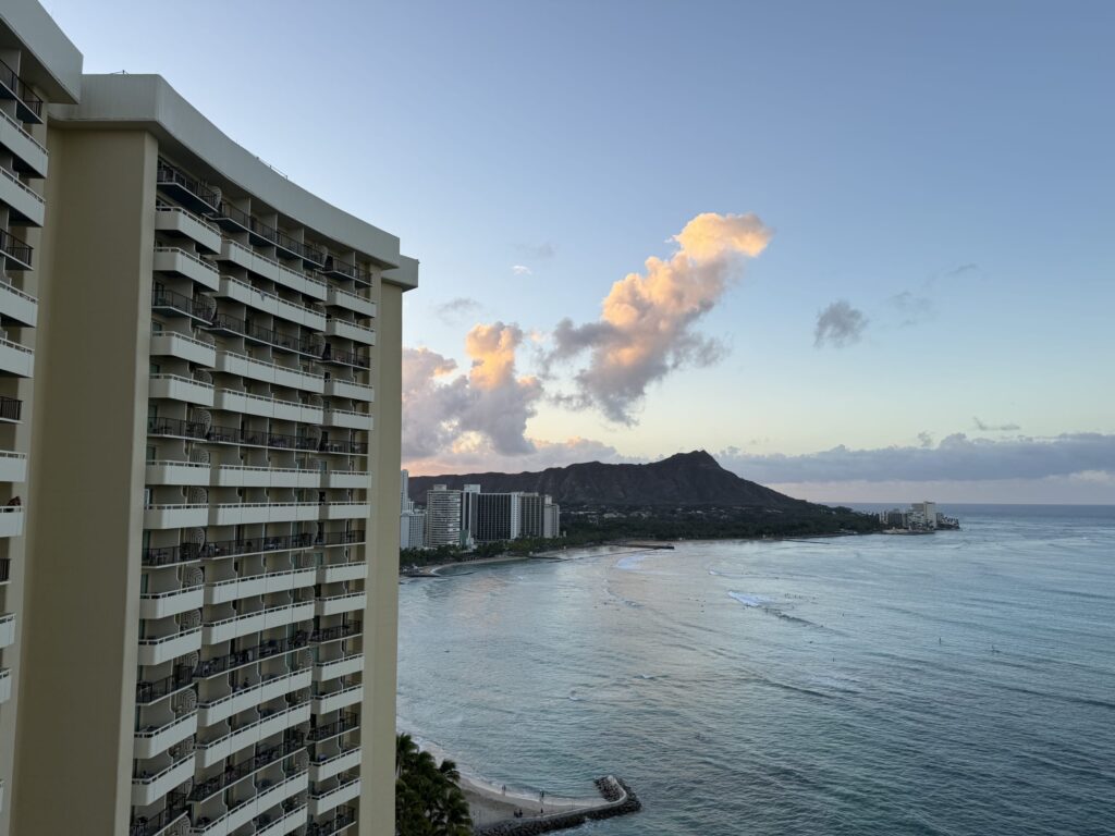 A view of Waikiki at dusk from a high floor of a hotel. Another hotel building is in the foreground, with Diamond Head and the ocean stretching out in the background.