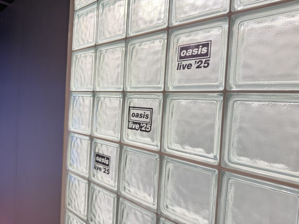斜めから撮影されたガラスブロックの壁。いくつかのブロックには、イギリスのロックバンドOasisのロゴと「live '25」という文字が書かれた黒いステッカーが貼られている。