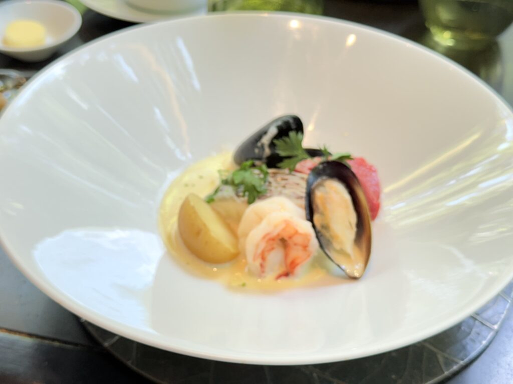 白い深皿の中央に盛り付けられた魚介料理。ムール貝、エビ、真鯛、ジャガイモがクリーム色のスープに入っている。