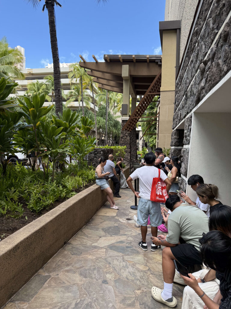 A line of people waiting on a flagstone pathway outside a building, surrounded by lush tropical plants and palm trees under a blue sky.