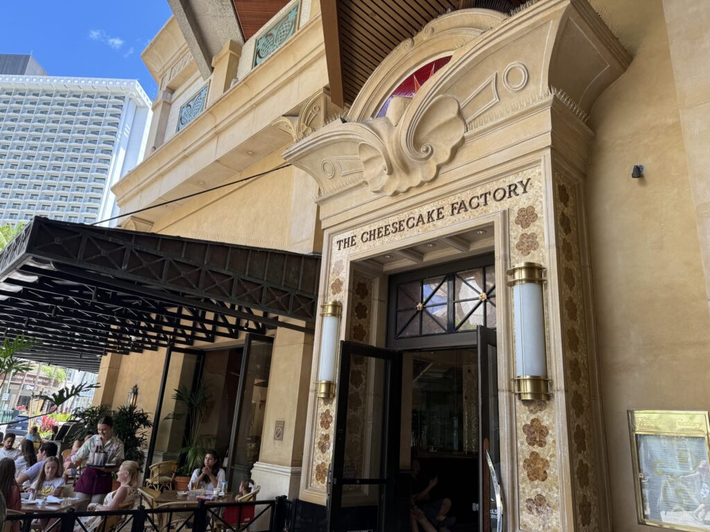 The ornate entrance of The Cheesecake Factory on a sunny day, with people enjoying their meals on the outdoor patio.
