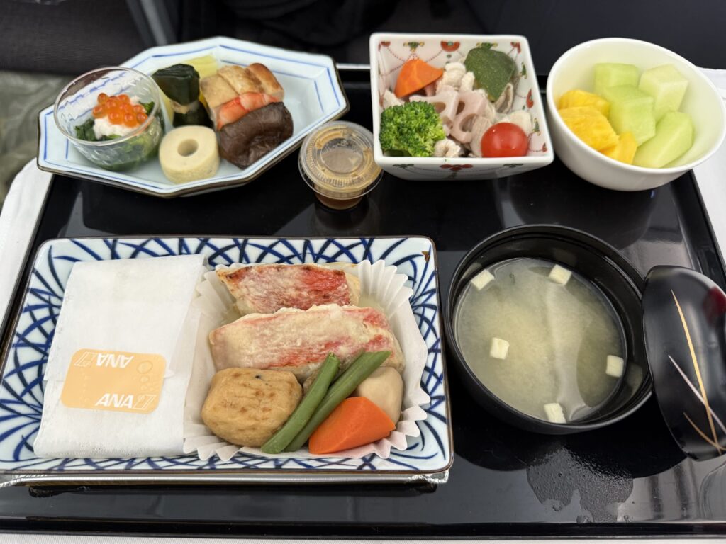 An ANA Japanese in-flight meal served on a black tray. It features a main dish of grilled fish, a small bowl with salmon roe, simmered vegetables, miso soup, and fresh fruit, all colorfully arranged.