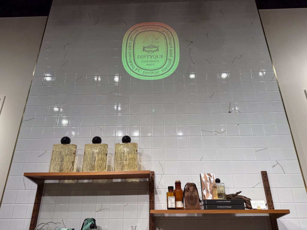 Inside a Diptyque store. Perfumes are arranged on wooden shelves against a white tiled wall, which features lines reminiscent of kintsugi and has the brand logo projected onto it.