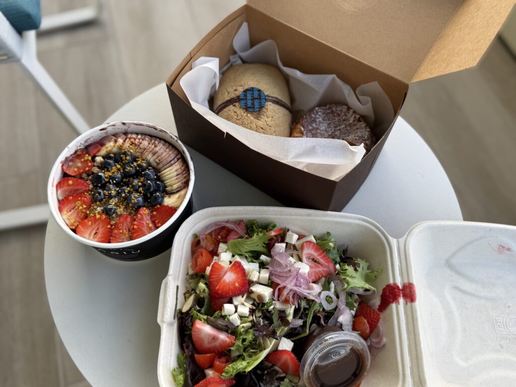 An acai bowl topped with fruit, a strawberry and feta salad, and a box with a cookie and pastry, all arranged on a white table.