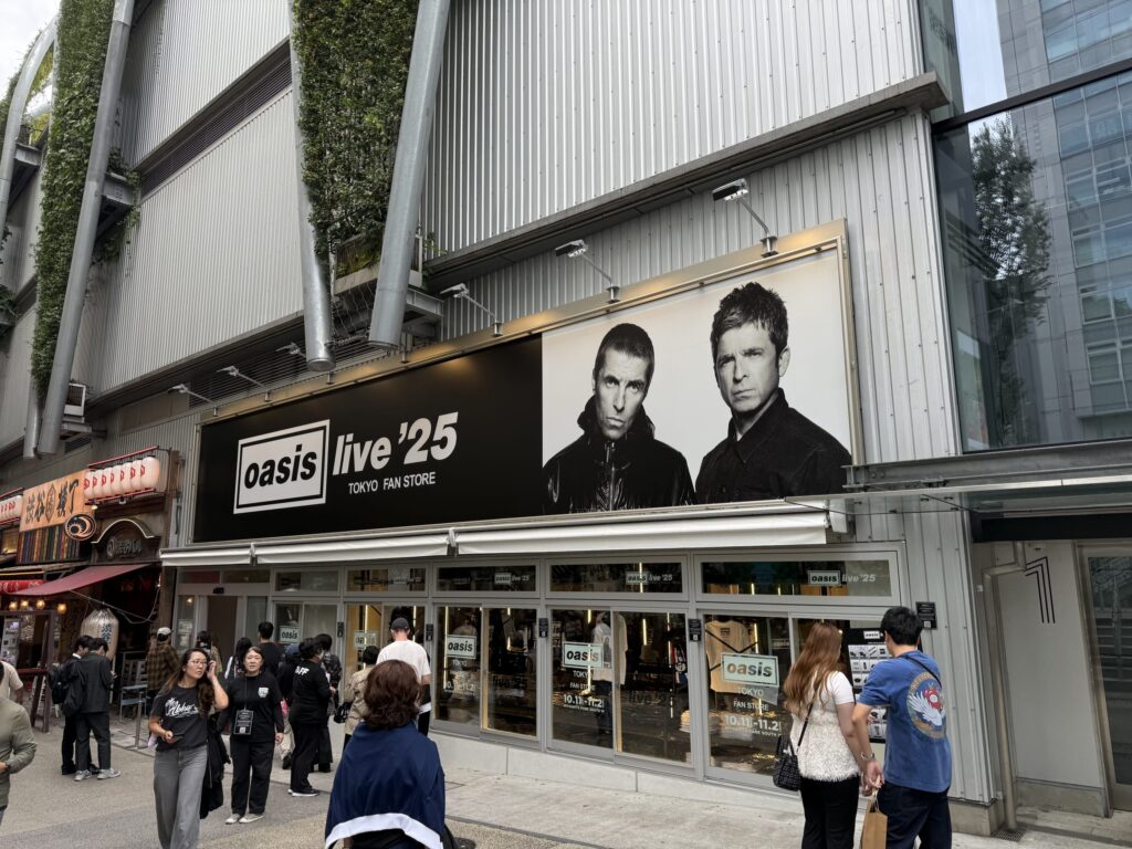 渋谷の街角にある「oasis live '25 TOKYO FAN STORE」のポップアップストアの外観。大きな看板にはリアム・ギャラガーとノエル・ギャラガーの白黒写真が掲げられ、店の前には多くのファンが集まっている。