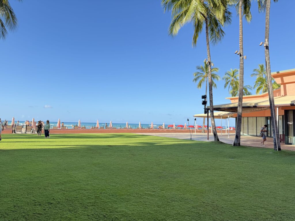 The courtyard of the Royal Hawaiian Hotel. Beyond a vast green lawn under a clear blue sky, Waikiki Beach and the ocean are visible with a line of pink umbrellas.