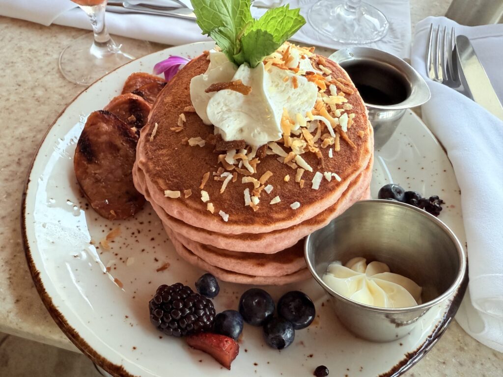 A plate of pink pancakes on a table at Surf Lanai in Hawaii. The stack of pancakes is topped with whipped cream, toasted coconut, and a mint sprig, with Portuguese sausage, fresh berries, butter, and syrup served on the side.