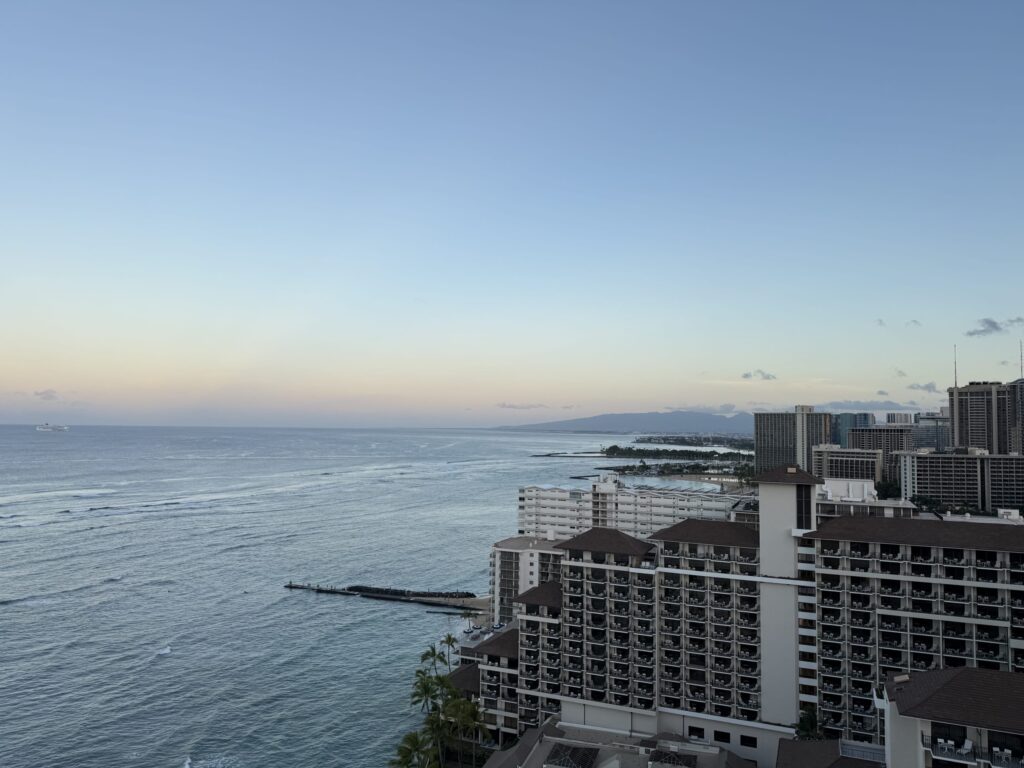 High-angle view of the Waikiki, Hawaii coastline at dawn. The blue Pacific Ocean is on the left, with large hotels like the Halekulani lining the shore on the right, and distant mountains under a pale blue and yellow sky.