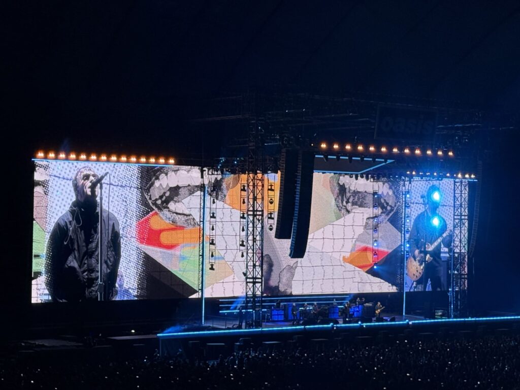 A concert stage at night. A giant screen shows singer Liam Gallagher at the microphone and a guitarist, while the band performs on the stage below. A large crowd is visible in the foreground.