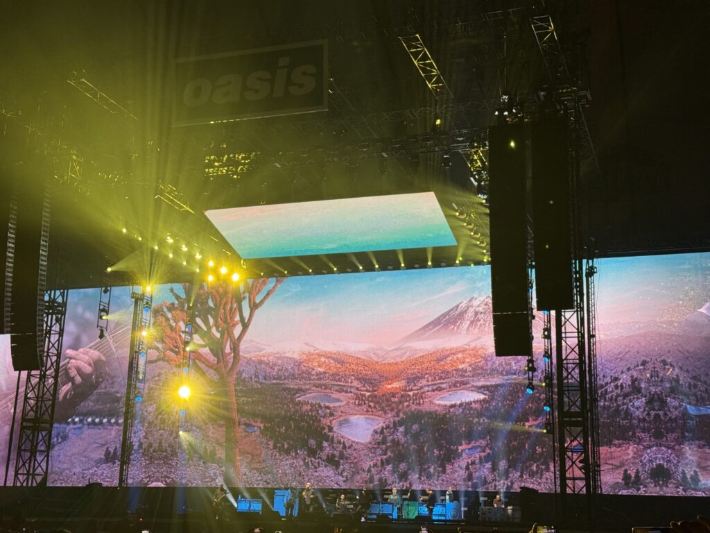 コンサートステージで演奏するバンド。背景の巨大スクリーンには富士山を含む雄大な風景が映し出され、上部には「oasis」のロゴが見える。