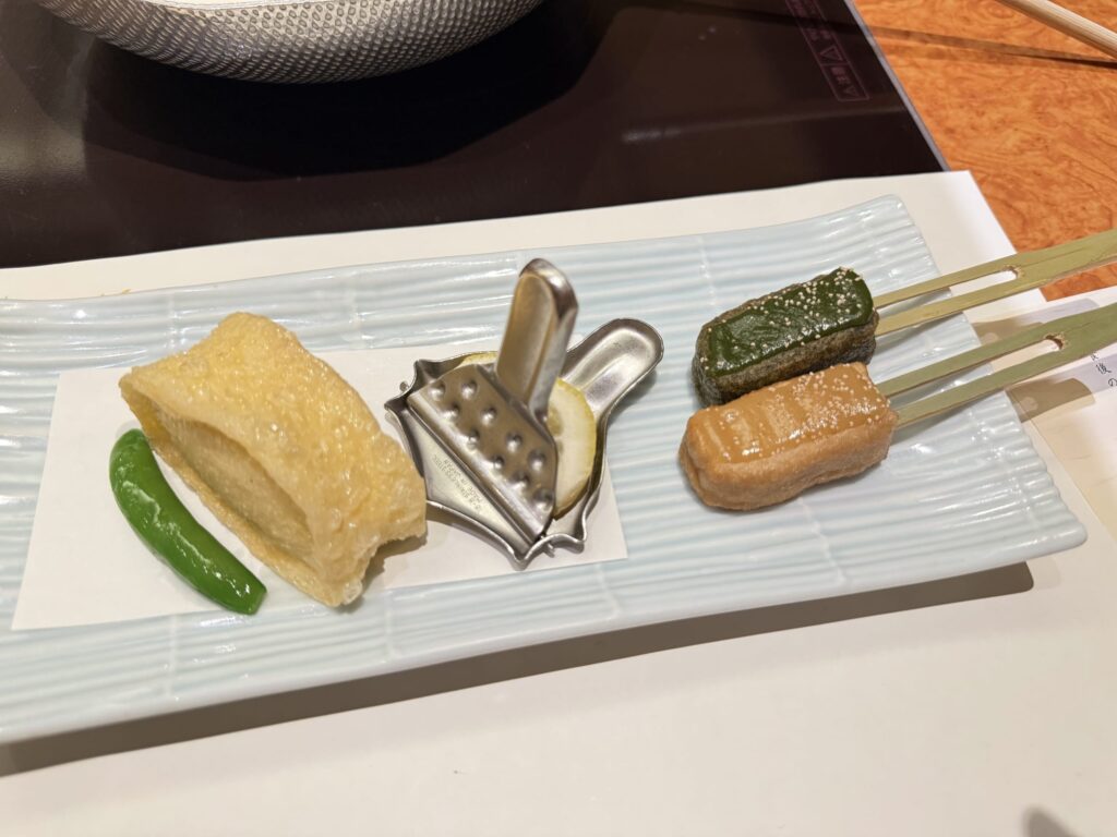 Yuba-age (fried tofu skin) and Nama-fu Dengaku (grilled gluten cakes with miso) arranged on a light blue rectangular plate. On the left are the yuba-age and a shishito pepper, on the right are two skewers of dengaku with green and light brown miso, and in the center is a lemon squeezer.
