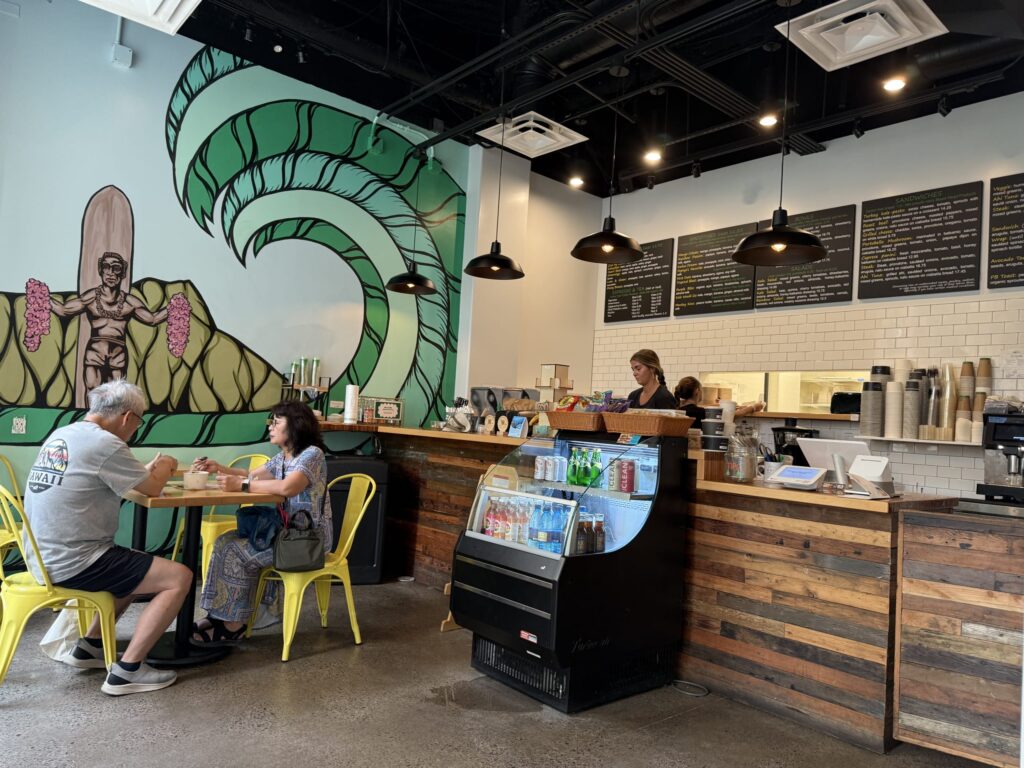 The interior of a Hawaiian-themed cafe. A large wave mural is painted on the wall, with customers sitting on yellow chairs and a wooden counter visible.