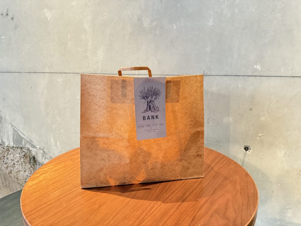 A brown paper bag with the 'BANK' logo on it, placed on a round wooden table against a bare concrete wall.