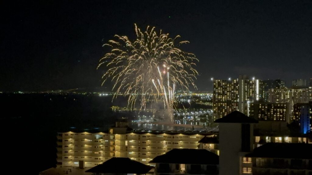 A large golden firework exploding over a coastal cityscape at night, with brightly lit buildings in the background.
