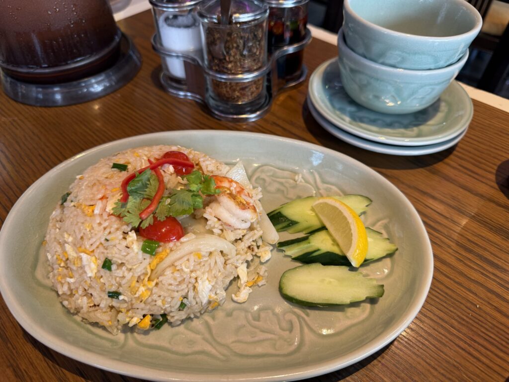 緑色の象柄の皿に盛られたタイ風のエビチャーハン。パクチーとカットトマトが乗っており、キュウリとレモンが添えられている。