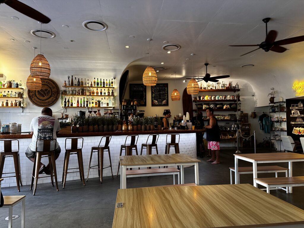 The interior of a Hawaiian cafe. A customer sits at the counter, with wooden tables in the foreground. Pineapples are lined up on the counter.