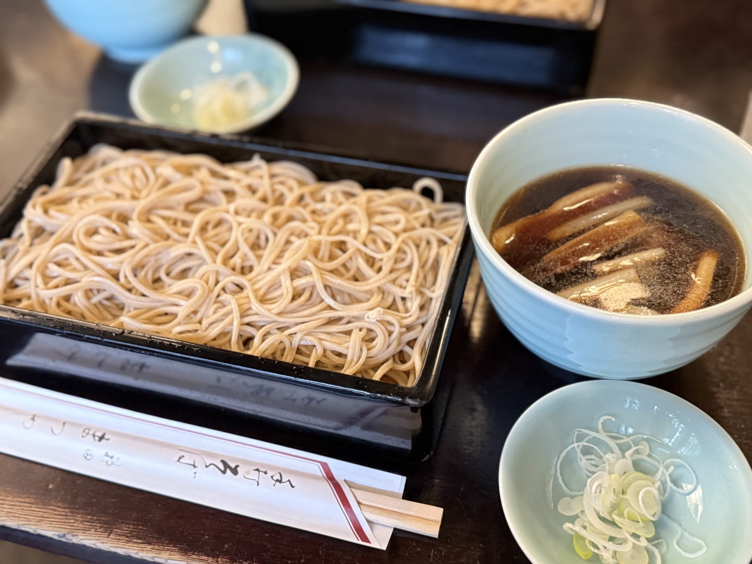 A Japanese meal set featuring cold soba noodles on a black seiro tray, a bowl of warm dipping sauce in a light blue bowl, and condiments like sliced green onions.