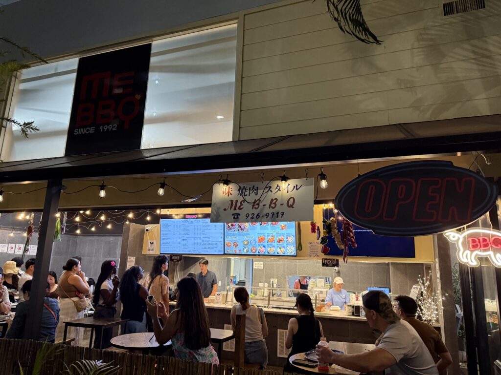 夜、焼肉レストラン「ME BBQ」の店先。カウンターには客が列をなし、テラス席も多くの人で賑わっている活気のある様子。