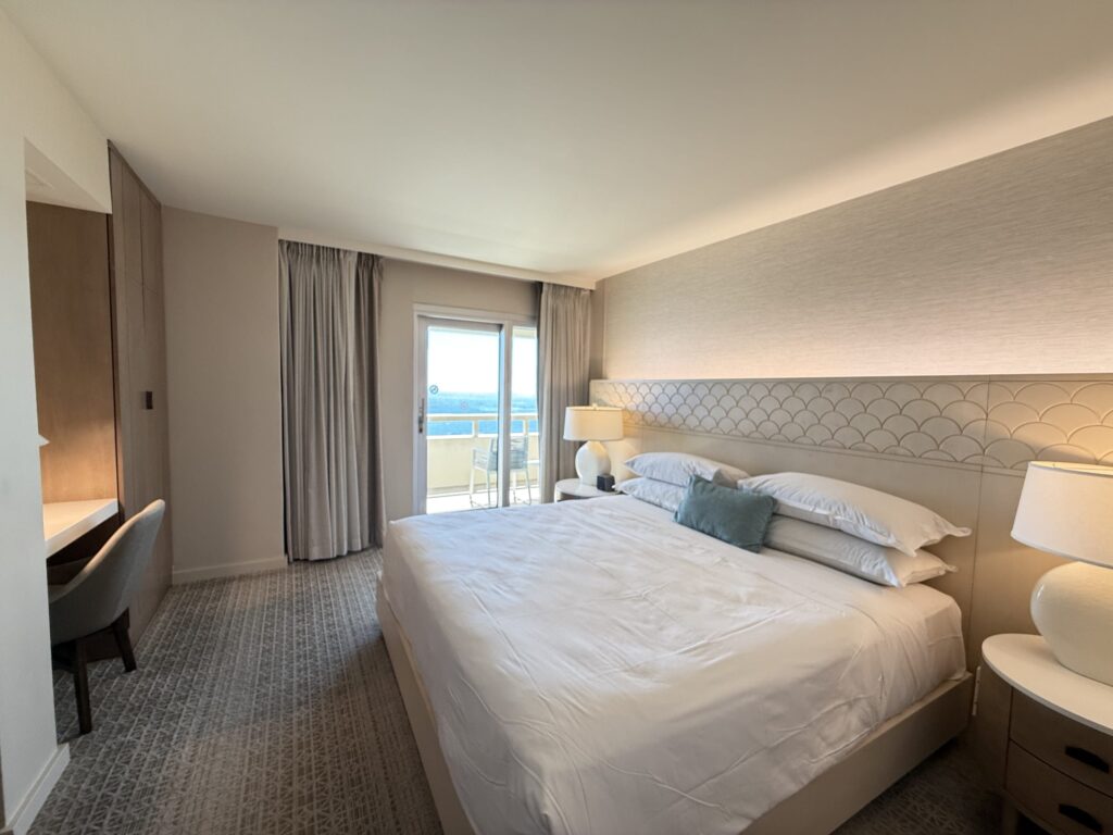 A hotel bedroom with a king-sized bed. In the background, there is a sliding glass door that opens to a balcony with an ocean view.