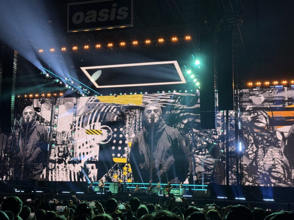 The stage at a Liam Gallagher concert. A giant screen shows a close-up of Liam's face with sunglasses as the band performs. The 'Oasis' logo is visible above the stage. The audience is enthusiastically holding up their smartphones.