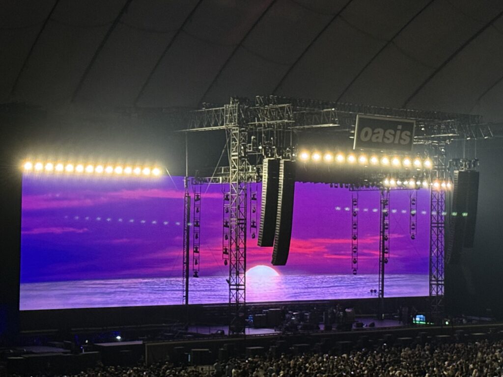 Oasisのコンサートステージ。巨大なスクリーンには紫とピンクの夕焼けが映し出され、大勢の観客がその光景を見上げている。