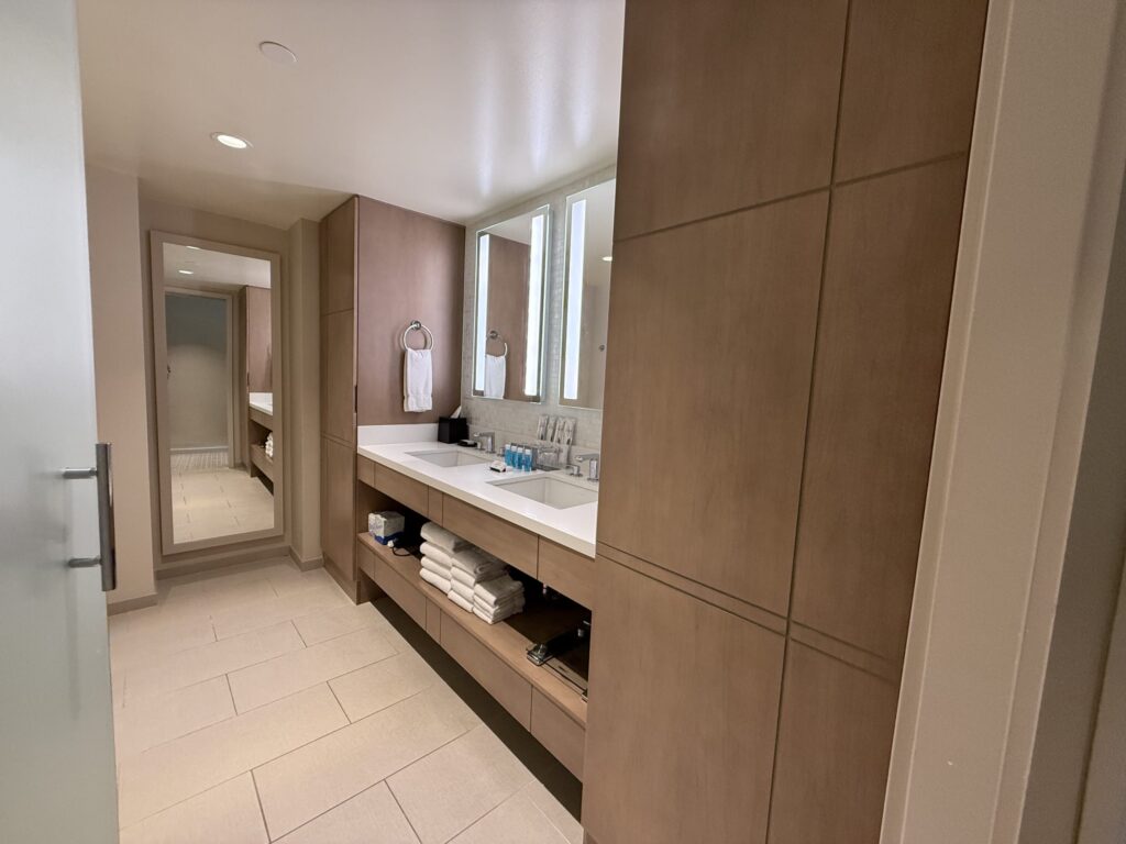 A modern hotel bathroom featuring a double vanity with wood-grain cabinets, a white countertop, two sinks, and two vertically lit mirrors.