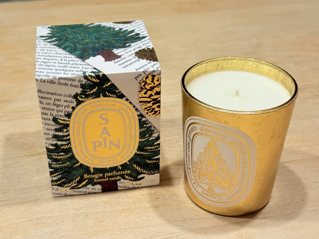 A diptyque Sapin scented candle in a gold vessel placed next to its decorative box with fir tree illustrations on a light wood surface.