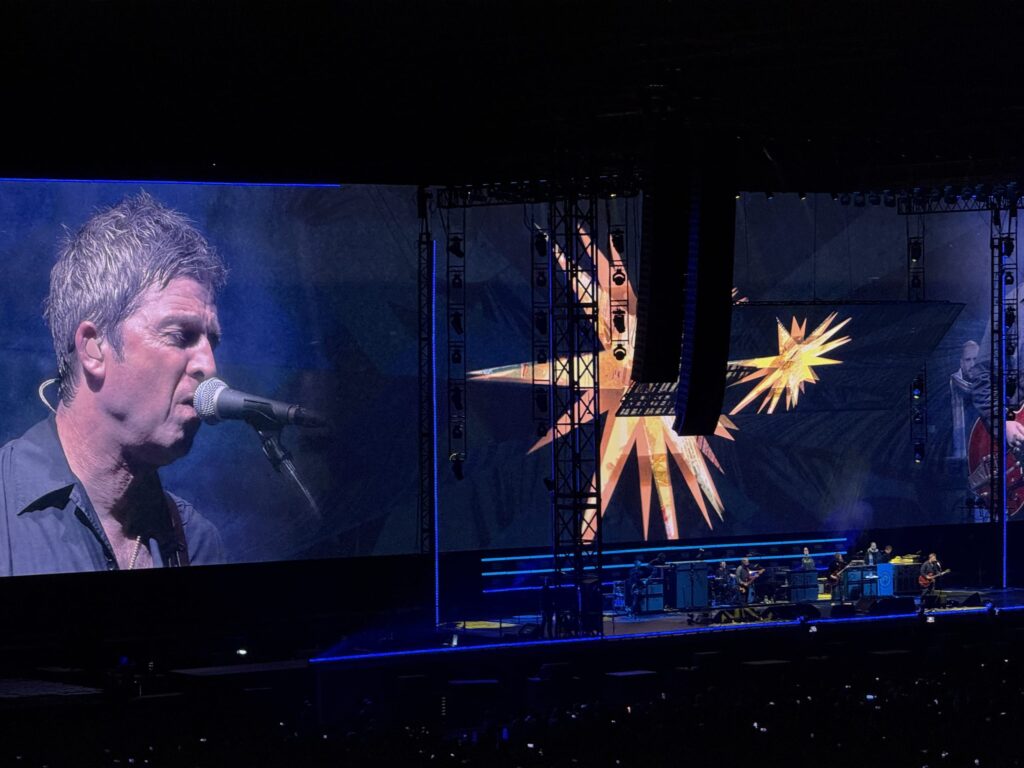 Noel Gallagher's High Flying Birds in concert. A large screen beside the stage shows a close-up of Noel Gallagher's face as he sings into a microphone, while the band performs on the stage.