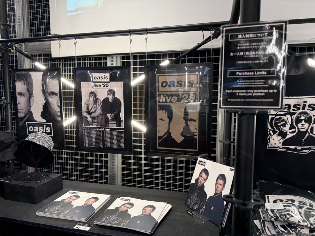 イギリスのロックバンド、オアシスのグッズ売り場の様子。壁にはギャラガー兄弟のポスターが飾られ、テーブルにはツアーパンフレットや帽子などのグッズが並んでいる。