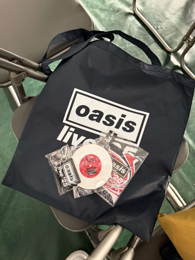 椅子の上に置かれた、Oasisのロゴ入り黒いトートバッグと、その上に乗せられたキーホルダー、ステッカー、レコード盤デザインのテープなどのVIP特典グッズ。