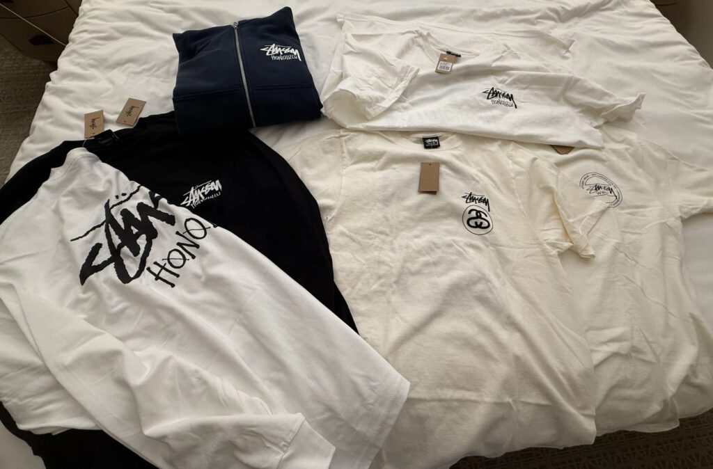 白いベッドの上に広げられた、Stüssyのホノルル限定Tシャツ、長袖Tシャツ、パーカーなどのアパレル商品。
