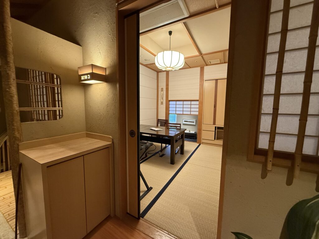 廊下から見える、温かい照明に照らされた和風の個室。部屋には畳が敷かれ、テーブルと椅子が置かれている。