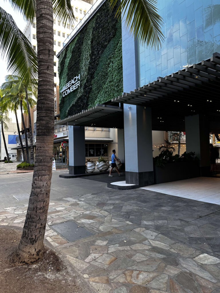 The entrance to the Waikiki Beachcomber hotel, featuring a striking green living wall, viewed from the sidewalk with a palm tree in the foreground.