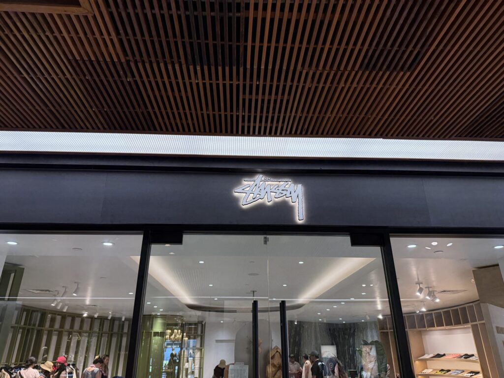 黒いファサードに白く光るロゴが特徴的なStüssyの店舗入り口。木製の格子天井の下、ガラス張りの店内には買い物客の姿が見える。