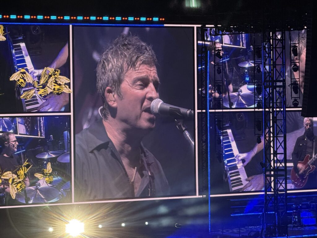 A giant screen on a concert stage. In the center, a large close-up of Noel Gallagher's face is shown as he sings into a microphone, surrounded by a collage of panels depicting his band members, including hands on a keyboard and a drummer.