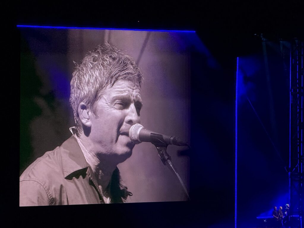 A black-and-white close-up of Noel Gallagher singing into a microphone is displayed on a giant screen on a concert stage. To the right, the actual stage illuminated by blue lighting is visible.