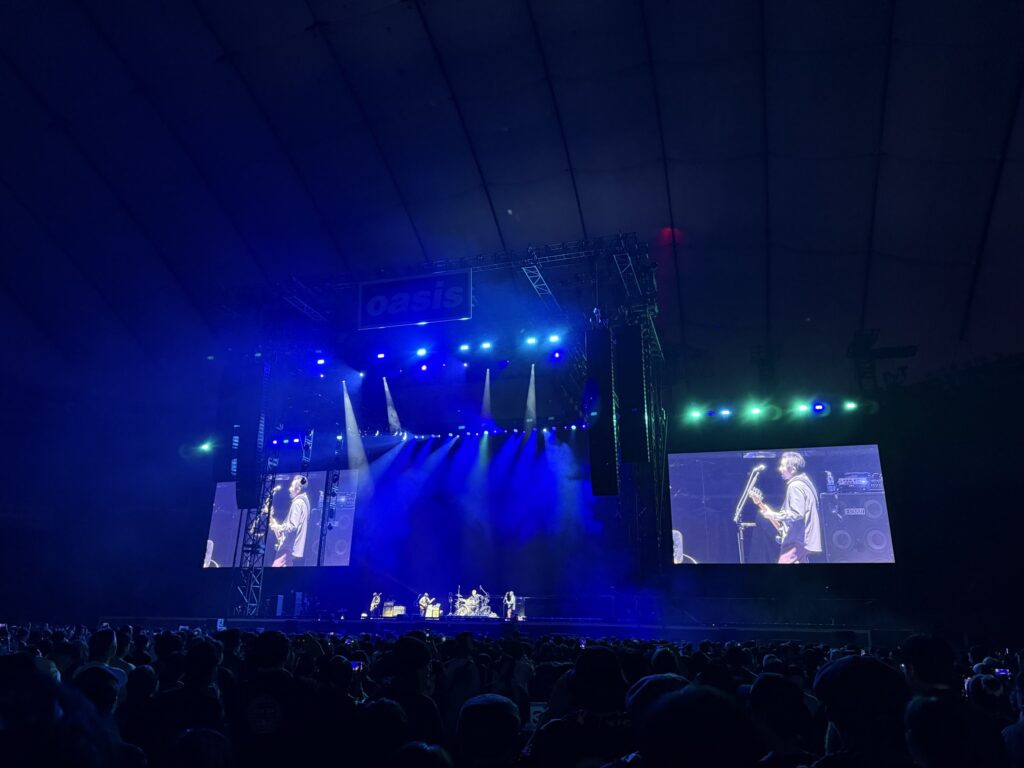 ASIAN KUNG-FU GENERATION performing on a stage in a dome venue bathed in blue light. Large screens on either side of the stage show the members, and silhouettes of a large crowd are visible in the foreground.