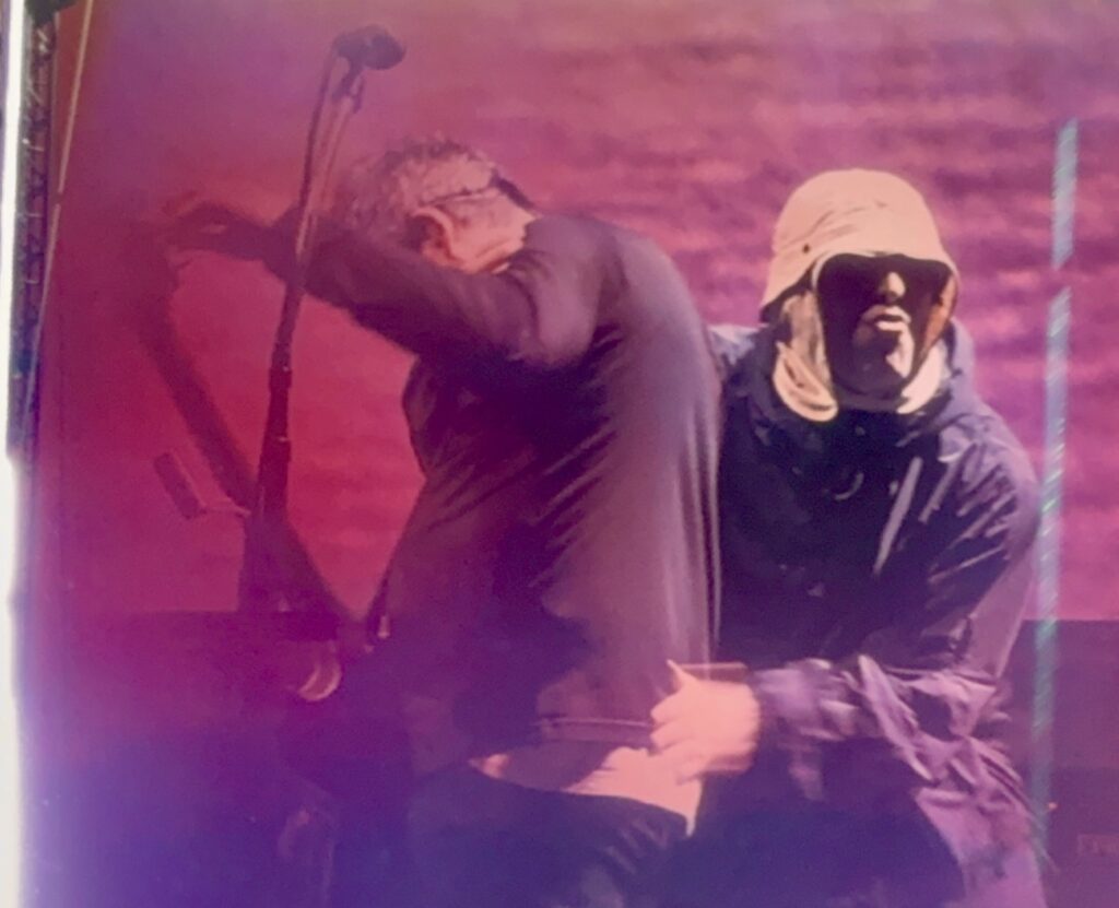 On a stage awash in pink light, a man in a hooded jacket hugs another man from behind, who is leaning into a microphone stand.