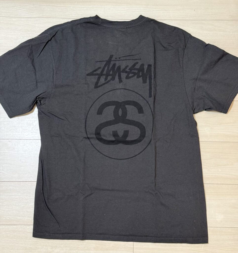 木の床に平置きされたステューシーの黒いTシャツ。背面には同系色の黒でストックロゴとSSリンクロゴがプリントされている。