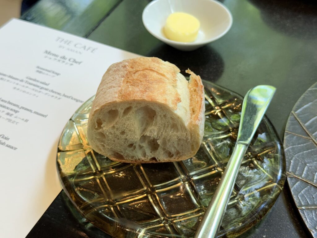 A piece of bread and a butter knife on a green glass plate. In the background, there is a small dish with butter and a restaurant menu.
