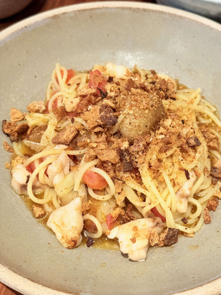 A close-up of monkfish and tomato spaghetti in a rustic gray bowl, topped with a generous amount of crispy breadcrumbs.