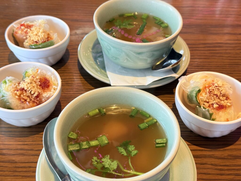 A lunch set for two on a wooden table. Each set consists of a light green bowl of soup and a small white bowl of fresh spring rolls.