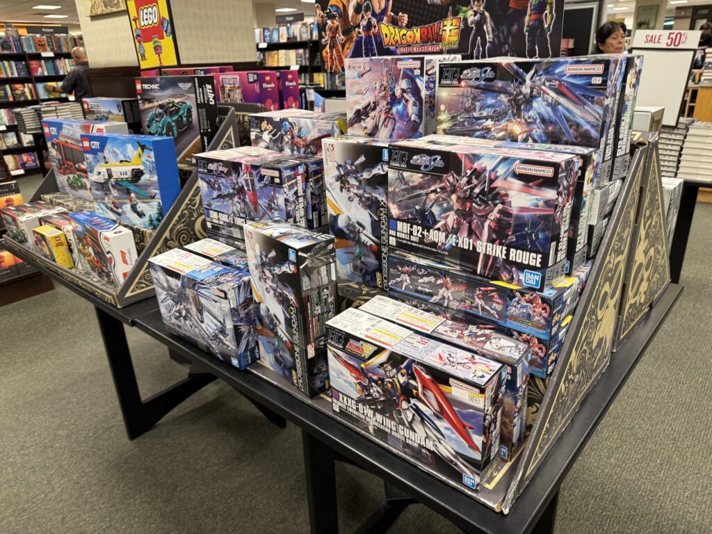 A display table inside a bookstore filled with various Gundam model kits (Gunpla) and LEGO sets, with bookshelves visible in the background.