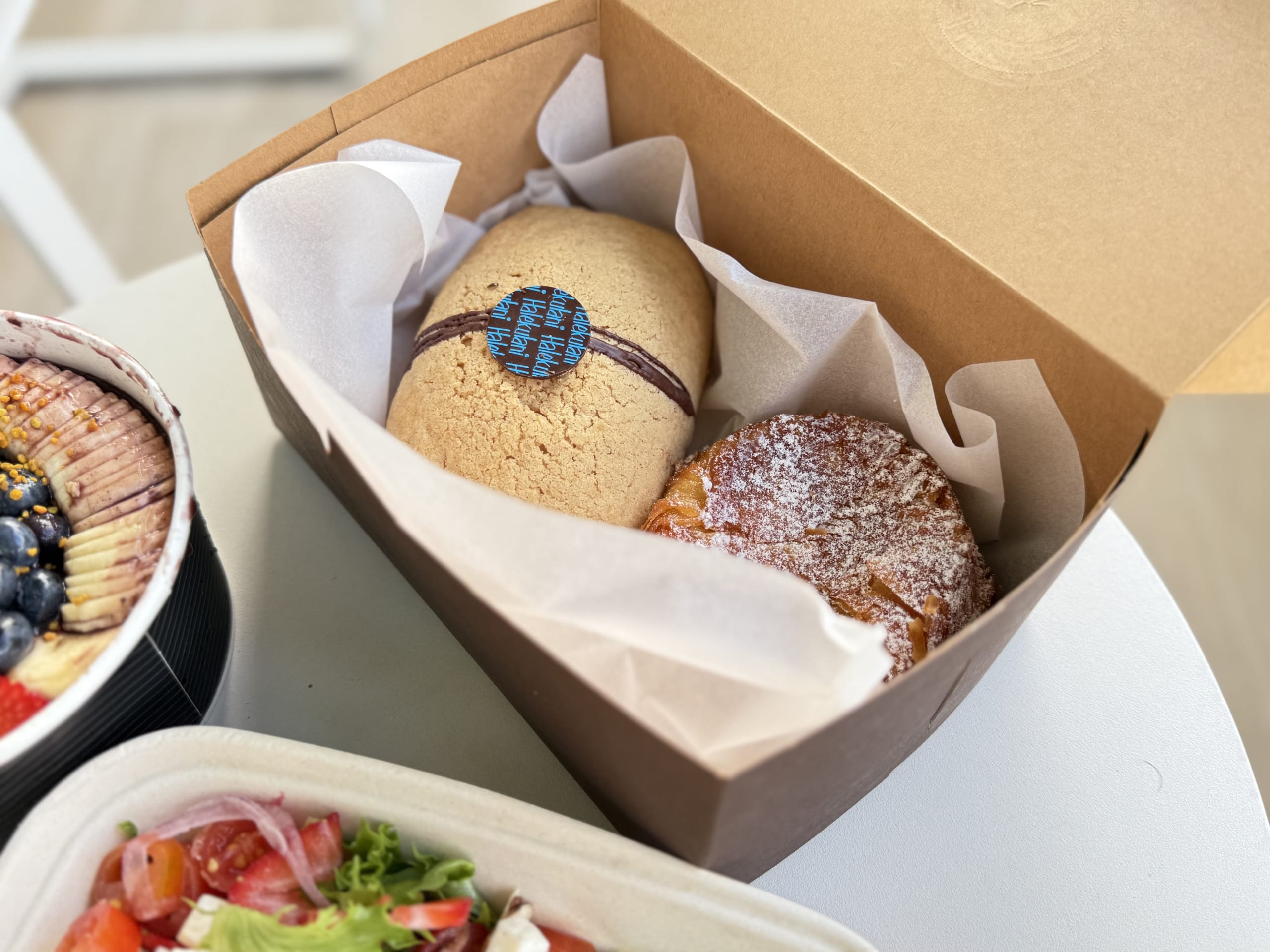 A takeout meal on a white table. An open box contains a cookie with the Halekulani logo and a pastry, placed next to an açaí bowl and a salad.